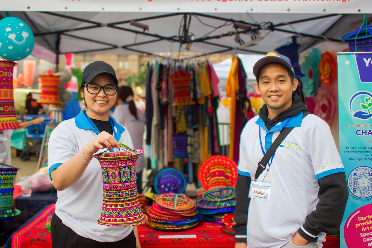 Two vendors smiling in a pop-up canopy booth full of woven crafts